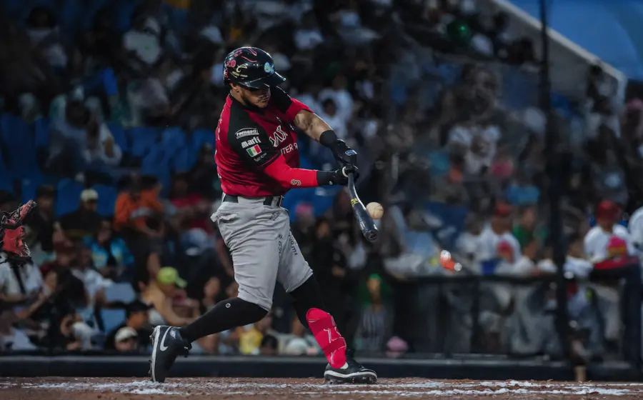 Bateador de los Guerreros de Oaxaca haciendo swing y conectando la pelota de béisbol durante un juego nocturno. Fotografía de acción profesional de la LMB con el jugador enfocado y el público desenfocado.