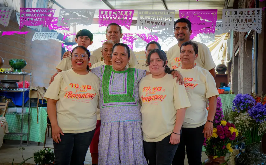 La chef Mimí López celebra sonriente en un evento gastronómico en Oaxaca, rodeada de su familia y el equipo de su cocina.