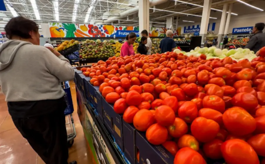 Una persona compra jitomates y papas en un mercado, ilustrando el aumento en el costo de la canasta básica que alcanza los 2,597 pesos.