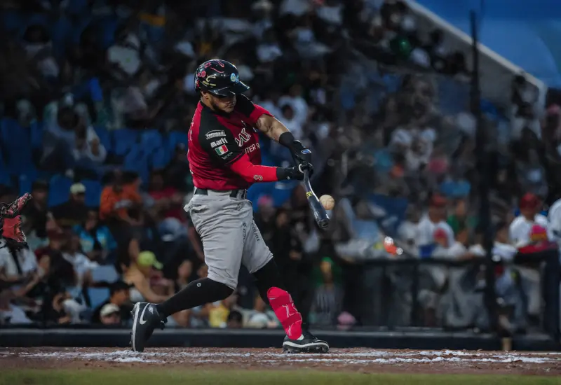 Bateador de los Guerreros de Oaxaca haciendo swing y conectando la pelota de béisbol durante un juego nocturno. Fotografía de acción profesional de la LMB con el jugador enfocado y el público desenfocado.