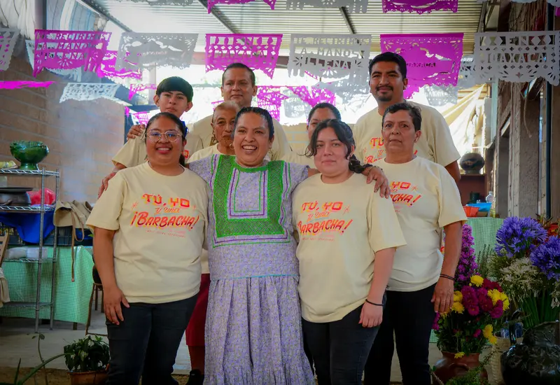 La chef Mimí López celebra sonriente en un evento gastronómico en Oaxaca, rodeada de su familia y el equipo de su cocina.