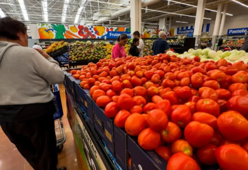 Una persona compra jitomates y papas en un mercado, ilustrando el aumento en el costo de la canasta básica que alcanza los 2,597 pesos.